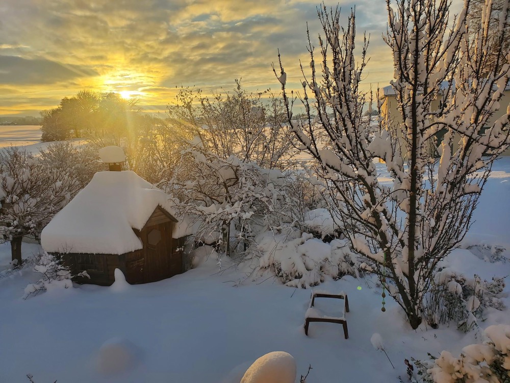 Blick über total verschneite Bäume und ein Gartenhaus, im Hintergrund ein orangefarbener Sonnenaufgang.